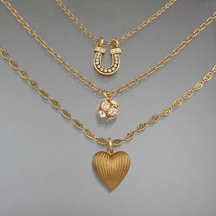 This is a charm necklace by John Wind / Maximal Art. Three chains, each with a different charm. Luck and Love themed with a horseshoe and a heart. Signed on the reverse of the horseshoe. Gold tone finish with rhinestones.  Approximately 5/8